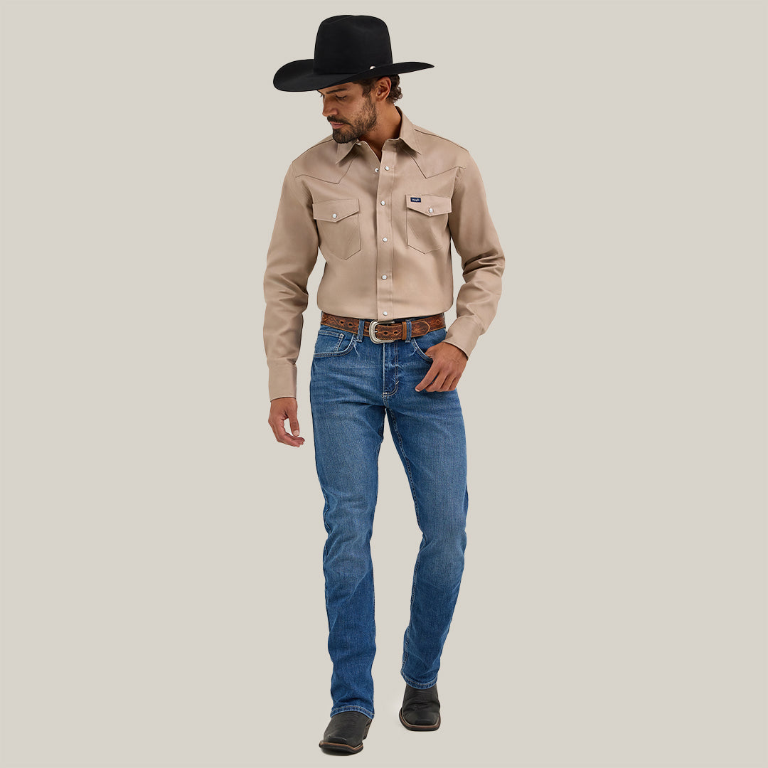 A man in a cowboy hat stands against a plain white background, wearing a beige long-sleeved shirt and Mens Wrangler 20X No. 42 Vintage Bootcut Jeans crafted from premium denim. Dark boots complete his rugged look as he gazes downward, embodying timeless Western style.