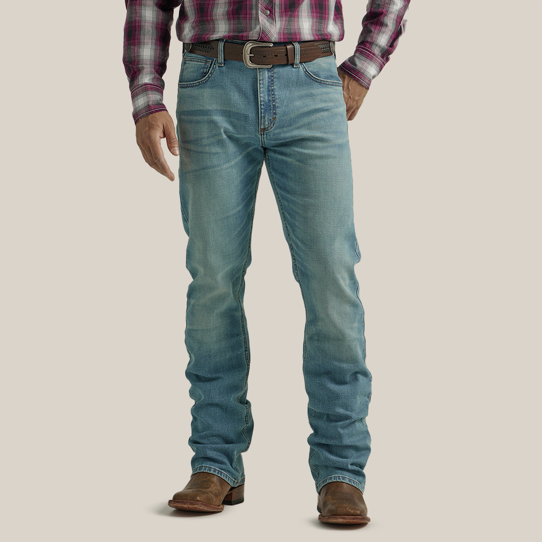 A person in Wrangler 20X Mens Light Wash Shade Bootcut Stretch Jeans and a plaid shirt stands facing away. The premium denim jeans boast decorative stitching on the back pockets, complemented by a brown belt and shoes, set against a plain, neutral backdrop.