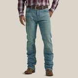 A person in Wrangler 20X Mens Light Wash Shade Bootcut Stretch Jeans and a plaid shirt stands facing away. The premium denim jeans boast decorative stitching on the back pockets, complemented by a brown belt and shoes, set against a plain, neutral backdrop.
