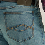 Someone in a plaid shirt, Wrangler 20X Mens Light Wash Shade Bootcut Stretch Jeans, and brown cowboy boots stands against a plain background. The light blue jeans are slightly faded and secured with a brown belt featuring an oval buckle.