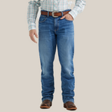 A person stands facing away wearing Wrangler 20x Mens Medium Wash Backwater Vintage Bootcut Stretch Denim Jeans with decorative stitching, a plaid shirt, and brown leather sandals against a light gray background.
