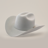 A light gray cowboy hat from the El Presidente collection, featuring a wide brim and slightly creased crown with a thin ribbon band. This 100X Independencia Platinum headwear rests on a neutral surface under subtle lighting.