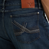 A person wears Wrangler 20X Sunnybrook Dark Wash Extreme Relaxed Straight Stretch Denim Jeans, paired with a gray shirt. The premium jeans feature decorative stitching on the back pocket and a brown leather patch with a small logo on the waistband.