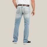 Rear view of a person in M7 Rocker Stretch Stirling Stackable Straight Leg Jeans - Shasta, displaying light blue faded stretch fabric. They wear a brown belt and white shirt, stand with one leg bent in black and white sneakers, emphasizing the relaxed straight-leg fit.