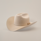 The 100X Independencia Belly, a luxury beige cowboy hat made from premium full beaver fur, boasts a curved brim and decorative band, elegantly showcased on a white surface.
