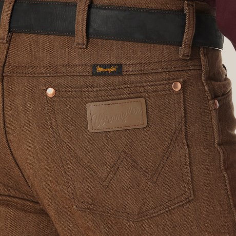 Close-up of a person wearing Wrangler Cowboy Cut Slim Fit Jeans - 100936BKW with a black belt. The jeans feature a branded leather patch above the right pocket and a small tag on the waistband, paired with a dark red shirt for classic style and rugged elegance.