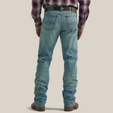 A close-up shows a person in Wrangler 20X Mens Light Wash Shade Bootcut Stretch Jeans (112335407) paired with a brown woven leather belt featuring a decorative round buckle, and a neatly tucked plaid shirt.