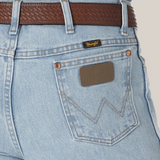 Close-up of a person in Wrangler Cowboy Cut Slim Fit Jeans - 0936GBH, light blue with a brown leather belt. The premium denim displays the classic W stitching on the back pocket with a leather Wrangler patch above it.